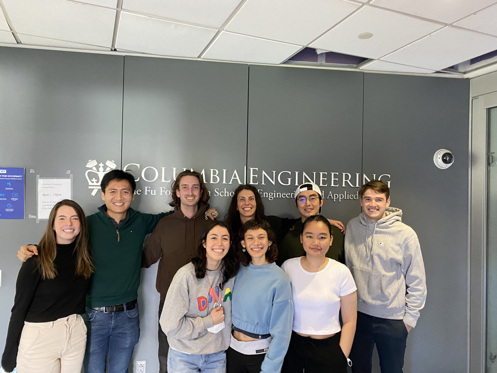 Nine researchers from the Marbella lab stand in a group in front of a Columbia Engineering logo
