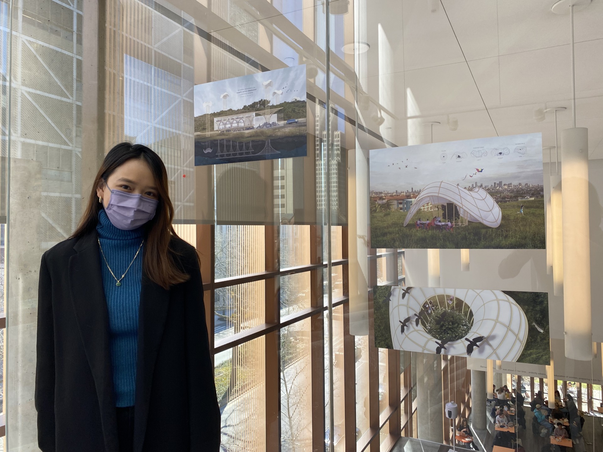 Shannon Hui wears a mask and stands beside three architectural renderings on the wall