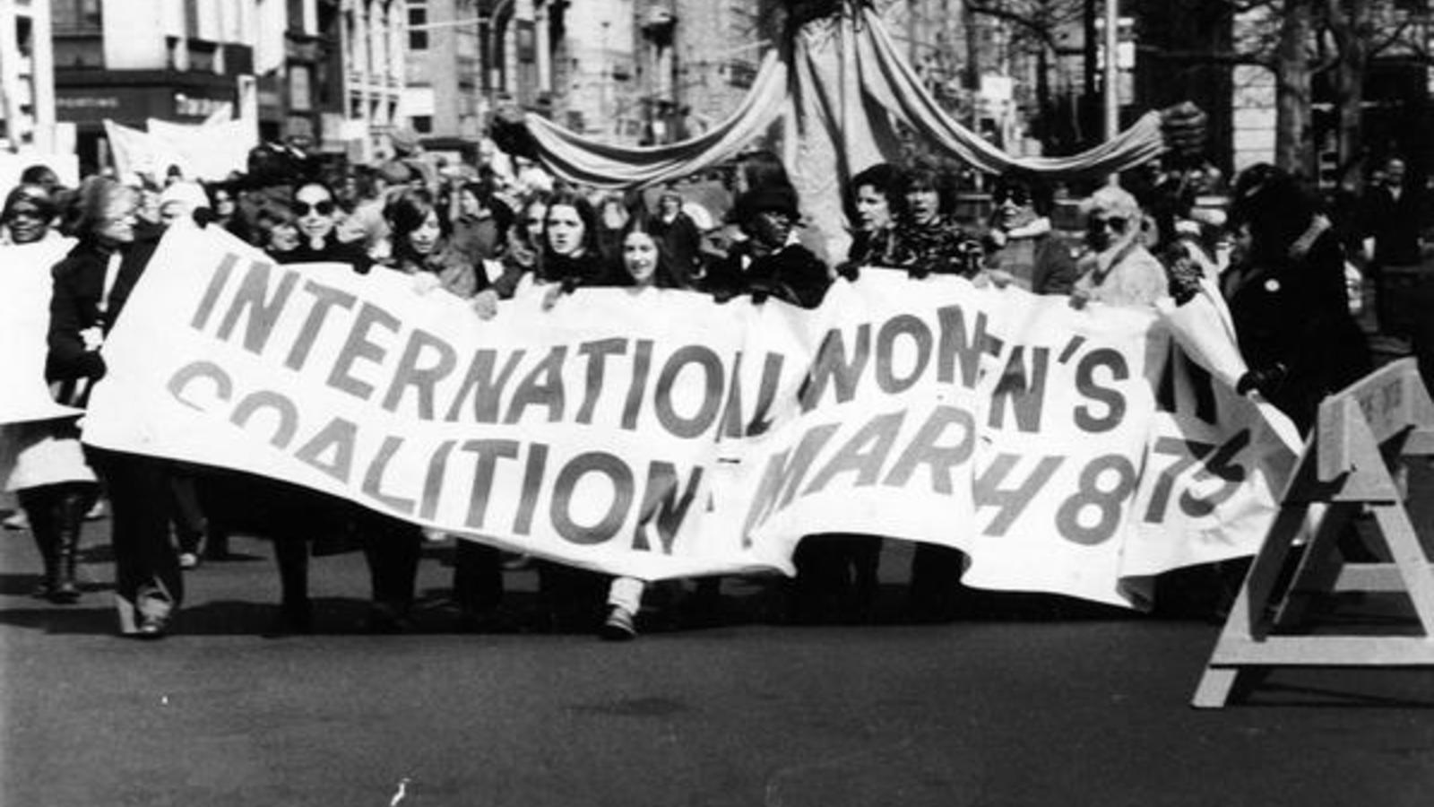 Marchers carrying banner that reads, "International Women's Coalition March, 8 1975." 